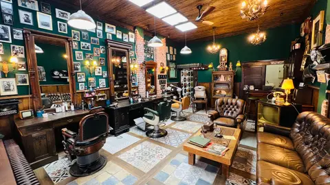 Der Laden vom Eingang aus: Barberstühle vor Holzspiegeln, gerahmte Porträts an grüner Wand, Chesterfield-Sofa